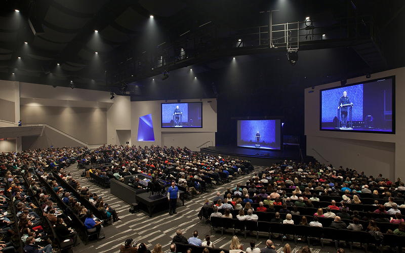 Speaker presenting in conference before large auditorium audience.